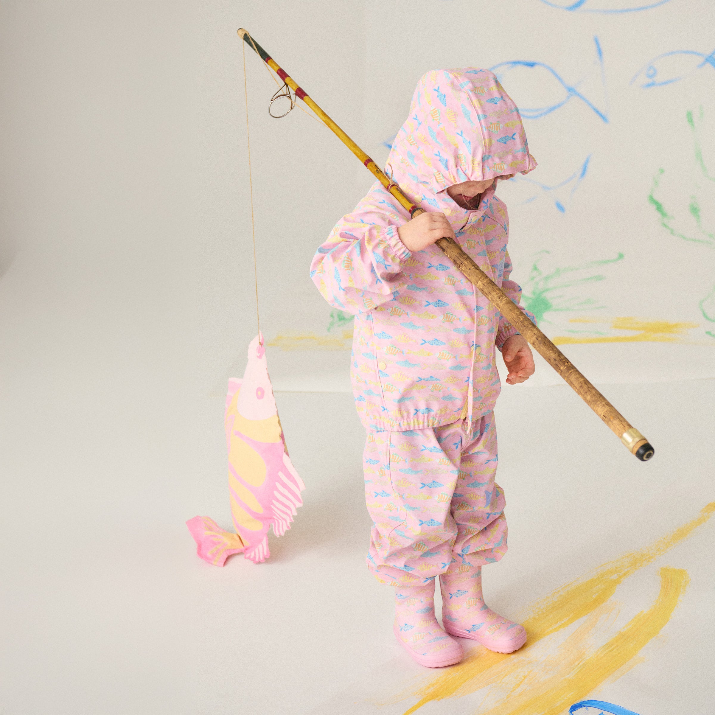 Barn iført Bundgaard x Helmstedt regntøj og gummistøvler i lyserødt fiskemønster, leger med en fiskestang. Farverigt og funktionelt regnsæt designet til leg og eventyr i al slags vejr.