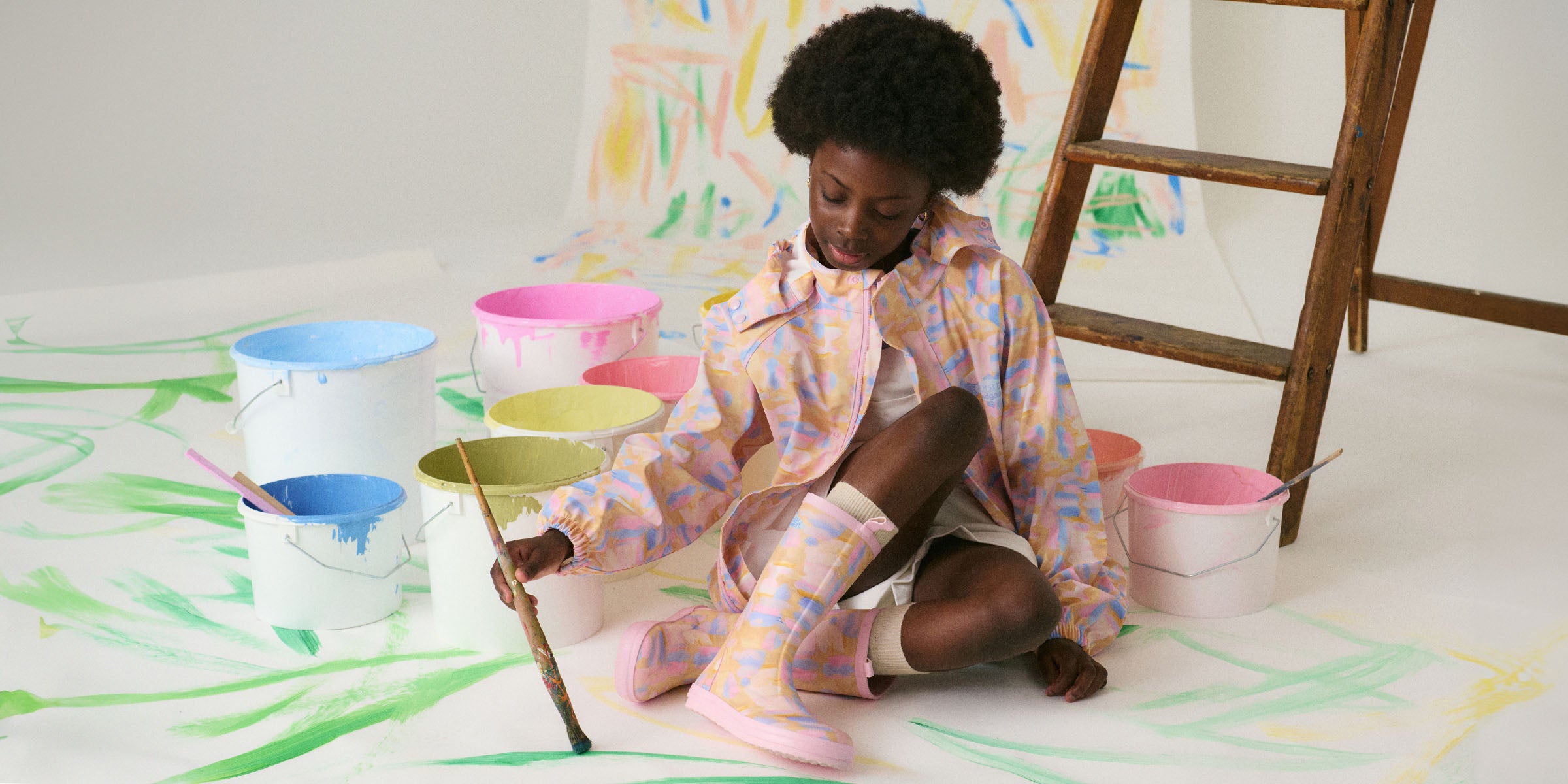 Barn iført Bundgaard x Helmstedt regnsæt og matchende gummistøvler i pastelfarver, maler kreativt omgivet af farverige malerbøtter. Stilfuldt og funktionelt regntøj til leg og eventyr.