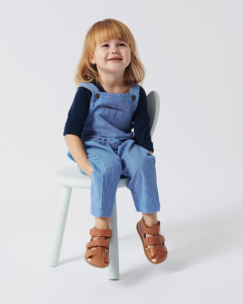 Smilende barn iført blåstribet overall og mørkeblå bluse, siddende på en hvid stol med komfortable, brune lædersandaler med velcrolukning. Perfekte sommersandaler til aktive børn.